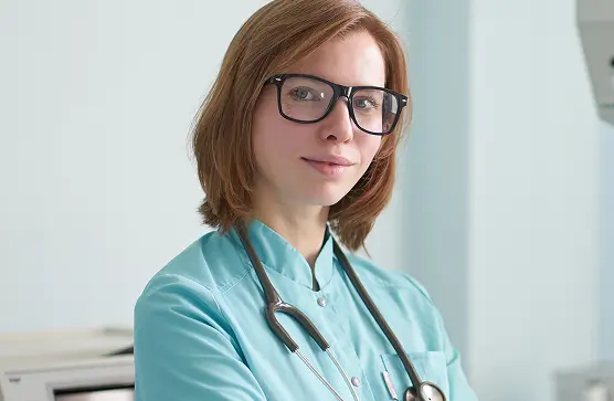 Junge Frau in medizinischer Arbeitskleidung mit Stethoskop steht in einem hellen Innenraum und blickt selbstbewusst in die Kamera. Die Szene vermittelt Professionalität im Gesundheitswesen.