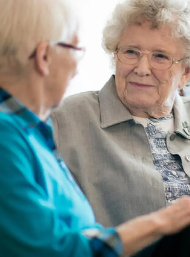 Zwei ältere Frauen sitzen nebeneinander und unterhalten sich. Eine der Frauen blickt die andere aufmerksam an. Die Szene vermittelt Gespräch, Nähe und soziale Begegnung im Alltag.