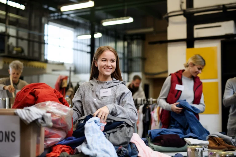 Eine junge Frau lächelt in die Kamera, während sie gemeinsam mit anderen Personen Kleidung sortiert. Die Szene spielt in einer großen Halle mit Kleiderspenden, im Hintergrund sind weitere Helferinnen und Helfer bei der Arbeit zu sehen.