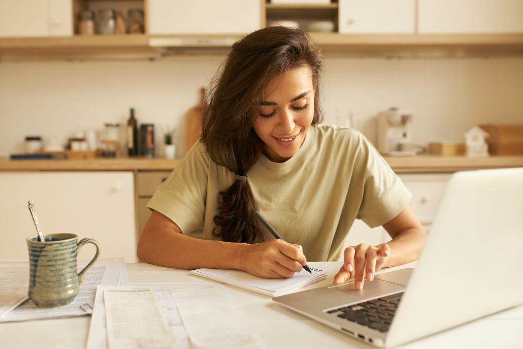Indoor image of student in t-shirt sitting at dining table in front of open laptop, studying distantly, drinking morning coffee and making notes in copybook, listening to lecture