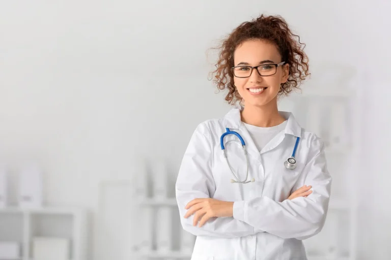 Junge Frau in medizinischer Arbeitskleidung mit Stethoskop lächelt in die Kamera. Sie steht vor hellem Hintergrund, die Szene vermittelt Professionalität im Gesundheitswesen.