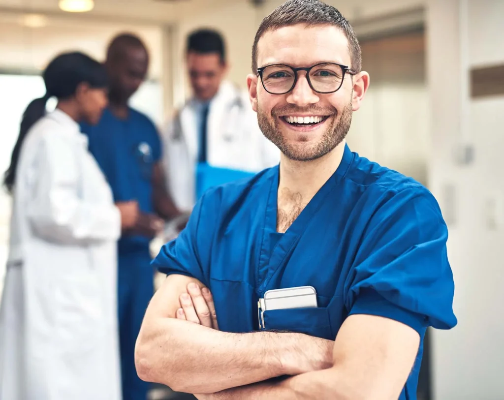 Mann in medizinischer Arbeitskleidung steht mit verschränkten Armen in einem Klinikflur und lächelt in die Kamera; im Hintergrund sind weitere medizinische Fachkräfte zu sehen.