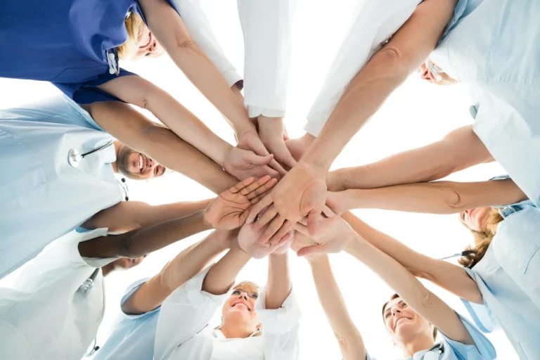 Mehrere Personen in medizinischer Kleidung legen ihre Hände übereinander als Zeichen von Teamarbeit und Zusammenhalt.