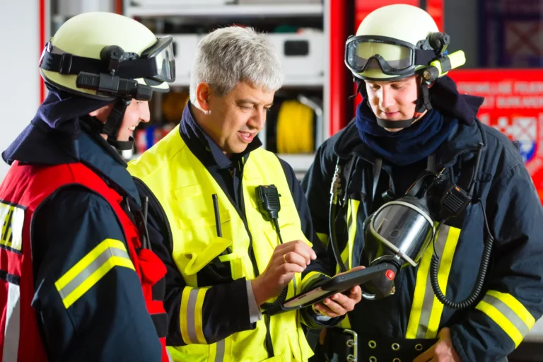 Drei Einsatzkräfte in Schutzkleidung und Helmen stehen zusammen und besprechen Einsatzdetails anhand eines Tablets.
