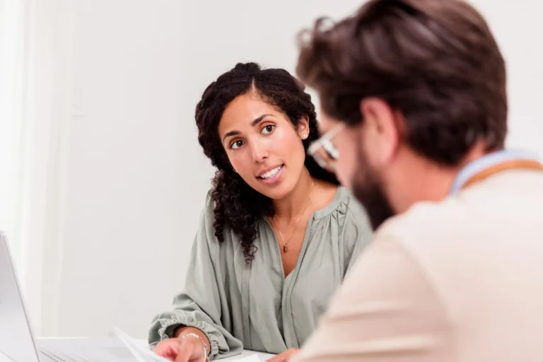 Zwei Erwachsene sitzen an einem Tisch und führen ein Gespräch. Eine Frau blickt aufmerksam und zugewandt zu einem Mann, der ihr gegenüber sitzt und Unterlagen vor sich hat. Die Frau gestikuliert leicht mit der Hand, was auf einen erklärenden oder beratenden Austausch hinweist. Die Szene wirkt ruhig und konzentriert und findet in einem hellen, neutralen Raum statt.