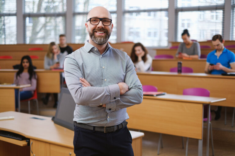 Mann steht mit verschränkten Armen in einem Hörsaal, im Hintergrund sitzen Studierende an Tischen.
