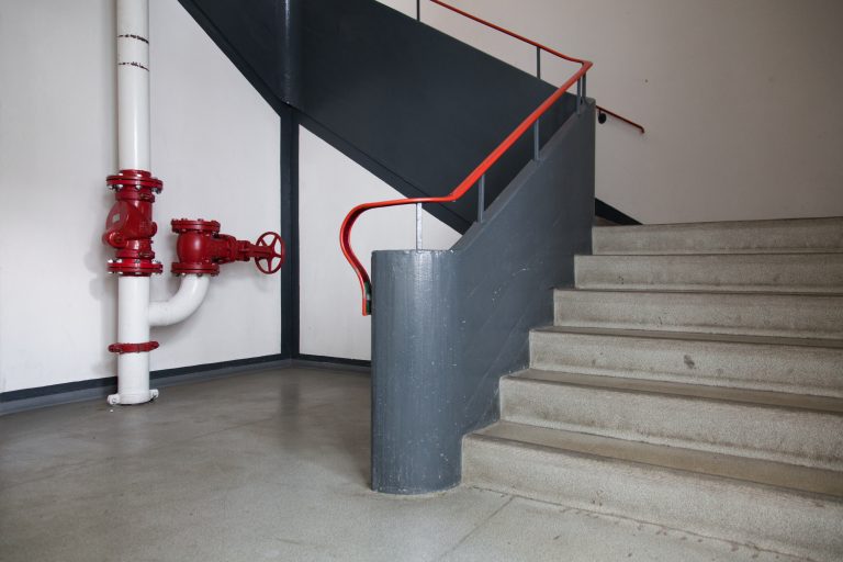 Treppenaufgang in einem Gebäude mit grauen Stufen, rotem Handlauf und sichtbaren Rohrleitungen an der Wand.