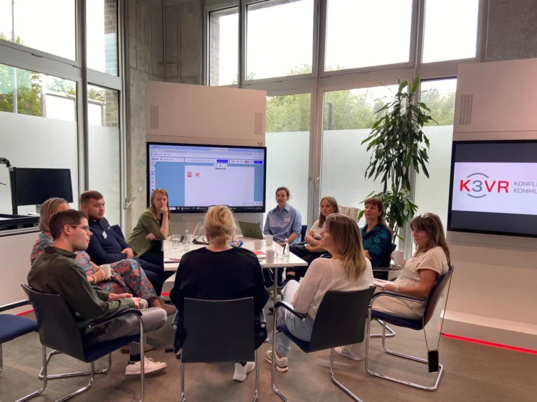 Mehrere Teilnehmende sitzen in einem Workshopformat im Kreis und diskutieren gemeinsam; im Hintergrund sind Präsentationsfolien auf einem Bildschirm zu sehen.