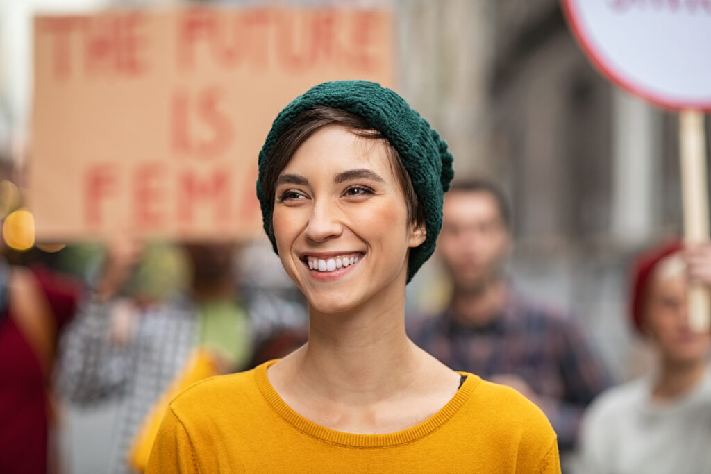 Eine junge Frau mit grüner Mütze und gelbem Pullover lächelt. Im Hintergrund sind Menschen mit Plakaten bei einer Demonstration zu sehen. Auf einem Plakat steht unscharf „The future is female“.