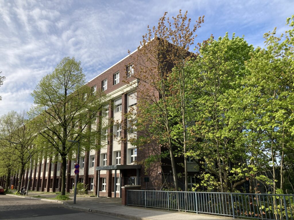 ChatGPT:Das Bild zeigt ein mehrstöckiges Backsteingebäude hinter einer Reihe grüner Laubbäume an einer ruhigen Straße unter leicht bewölktem Himmel.