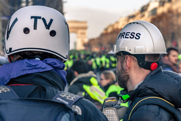 Zwei Pressevertreter mit Helmen, auf denen „TV“ und „PRESS“ steht, beobachten eine Demonstration. Im Hintergrund sind viele Menschen in gelben Westen sowie der Arc de Triomphe zu sehe