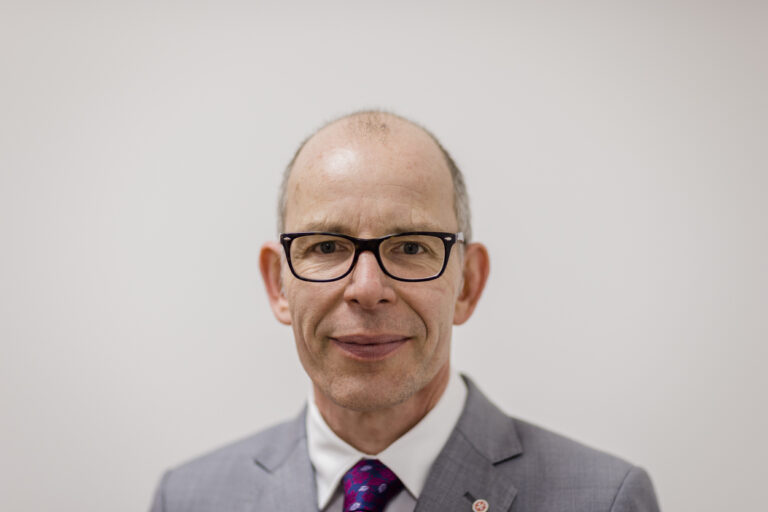 Porträt eines Mannes mit Brille in grauem Anzug und Krawatte vor neutralem, hellem Hintergrund.