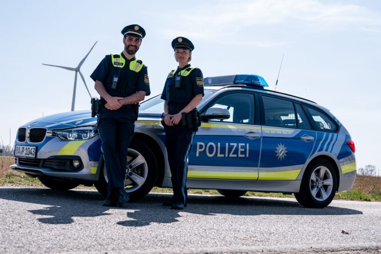 Zwei uniformierte Polizeikräfte stehen nebeneinander vor einem Polizeifahrzeug auf einer Landstraße. Beide tragen Schutzwesten, Dienstmützen und Funkgeräte. Hinter ihnen steht ein blau-silberner Streifenwagen mit eingeschaltetem Blaulichtbalken. Im Hintergrund sind Windräder und eine offene Landschaft zu sehen.