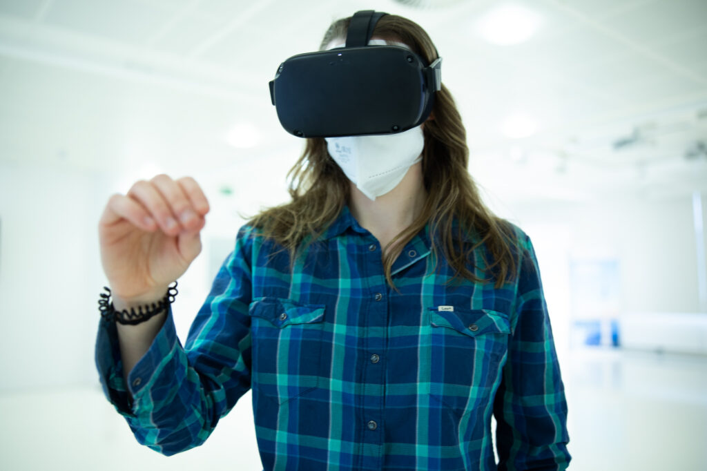 Person trägt eine VR-Brille und eine Schutzmaske und interagiert mit virtuellen Elementen in einem hellen Raum.