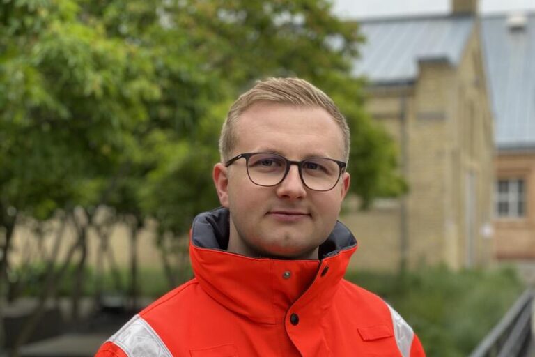 Porträt eines jungen Rettungskräfters in orangefarbener Einsatzjacke. Er trägt eine Brille, schaut direkt in die Kamera und steht im Freien vor Bäumen und einem Gebäude.