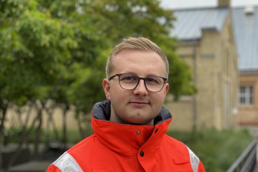 Porträt eines jungen Rettungskräfters in orangefarbener Einsatzjacke. Er trägt eine Brille, schaut direkt in die Kamera und steht im Freien vor Bäumen und einem Gebäude.