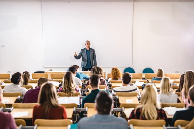 Ein Dozent steht vor einer Gruppe Studierender in einem Hörsaal und hält eine Vorlesung. Die Studierenden sitzen in Reihen und hören aufmerksam zu, im Hintergrund ist eine große weiße Projektionsfläche zu sehen.