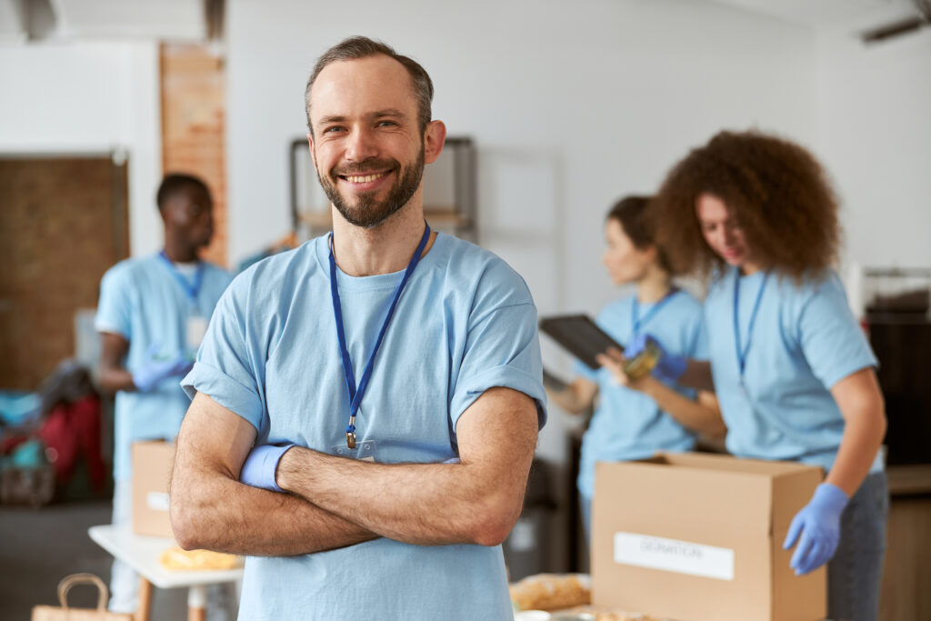 Freiwillige Helferinnen und Helfer in blauen T-Shirts sortieren Spenden und Lebensmittel, im Vordergrund lächelt ein Mann in die Kamera.
