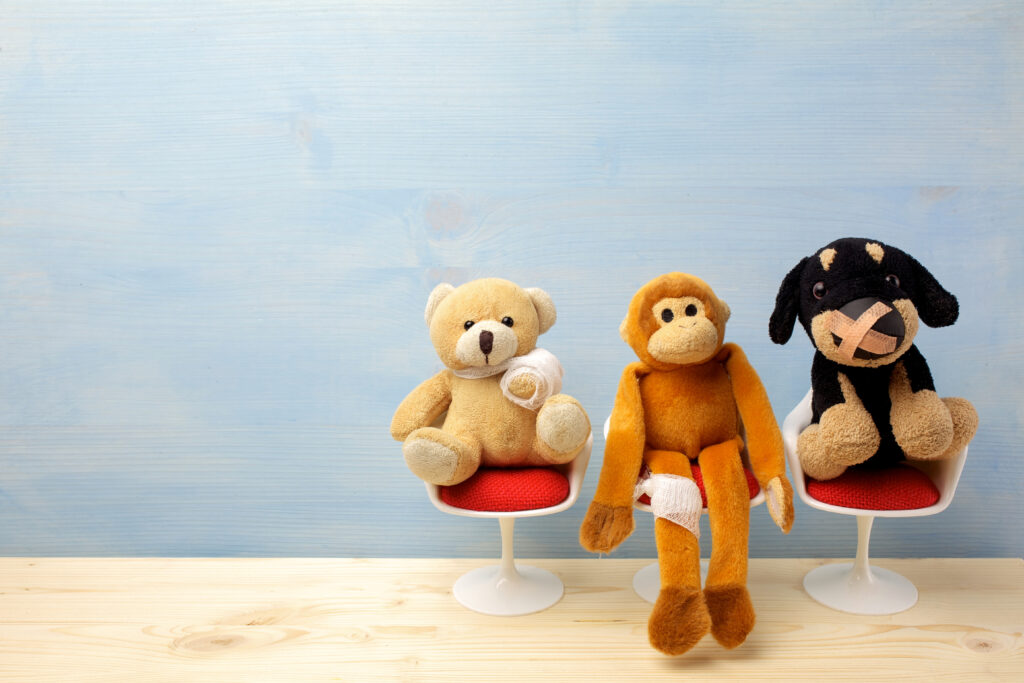 Drei Stofftiere (Teddybär, Affe und Hund) sitzen mit Verbänden auf kleinen Stühlen in einem hellen Raum.