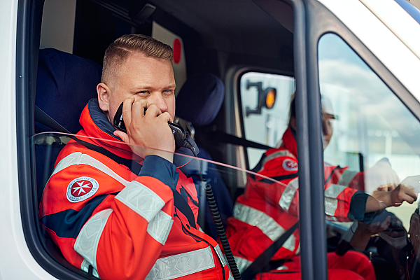 Ein Mitarbeiter aus dem Rettungsdienst fährt mit seiner Kollegin im Rettungswagen und spricht in ein Funkgerät.