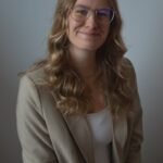 Porträt einer Frau mit langen, leicht gewellten Haaren und Brille. Sie trägt einen hellen Blazer über einem dunklen Oberteil und blickt freundlich in die Kamera. Neutraler Hintergrund.