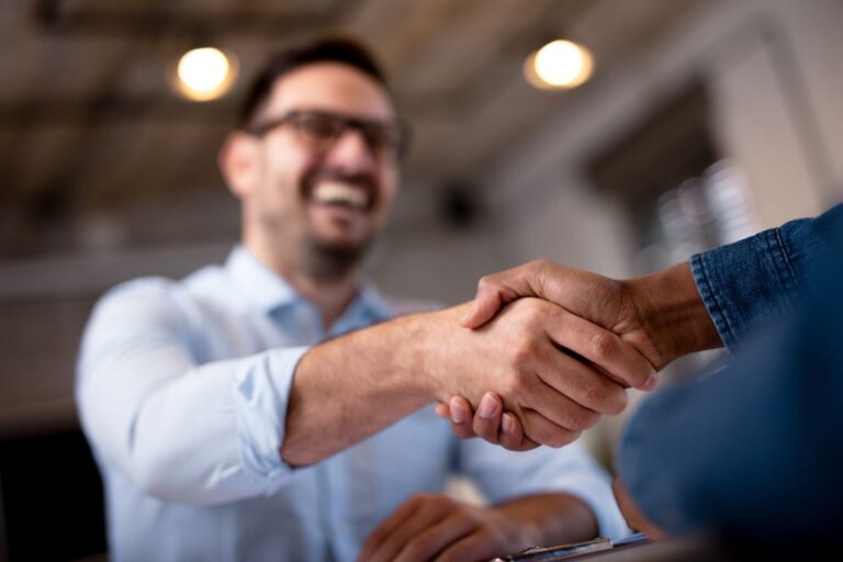 Zwei Personen geben sich die Hand in einem modernen Innenraum. Eine Person im Vordergrund trägt ein blaues Hemd, die andere ist im Hintergrund leicht unscharf und lächelt. Die Szene vermittelt einen professionellen und freundlichen Austausch.