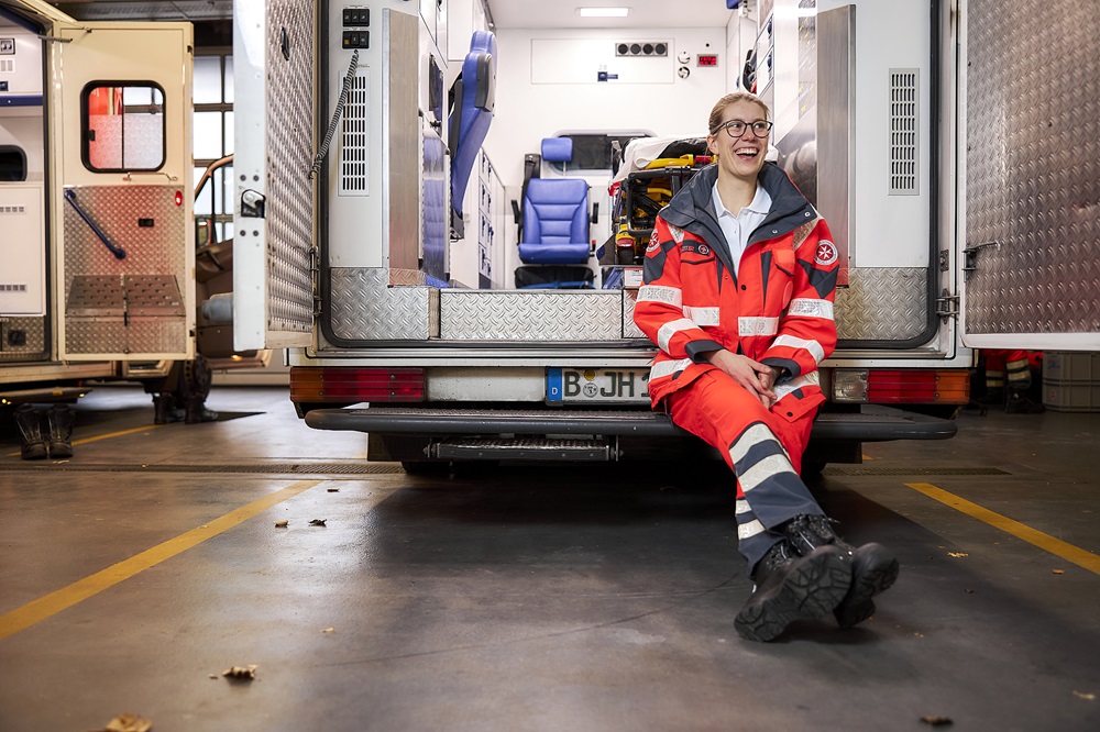 Rettungssanitäter in roter Einsatzkleidung sitzt vor einem Rettungswagen in einer Fahrzeughalle.