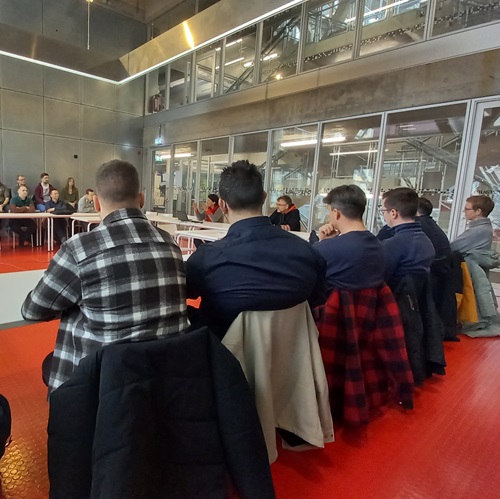 Studierende sitzen an einem langen Konferenztisch in einem modernen Gebäude mit rotem Teppichboden.
