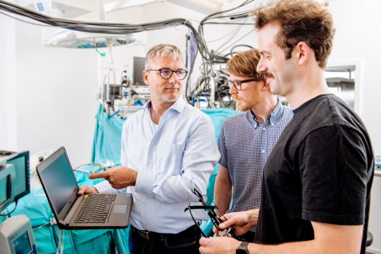 Drei Personen stehen in einem klinischen Umfeld und besprechen technische Inhalte anhand eines Laptops und medizinischer Geräte.