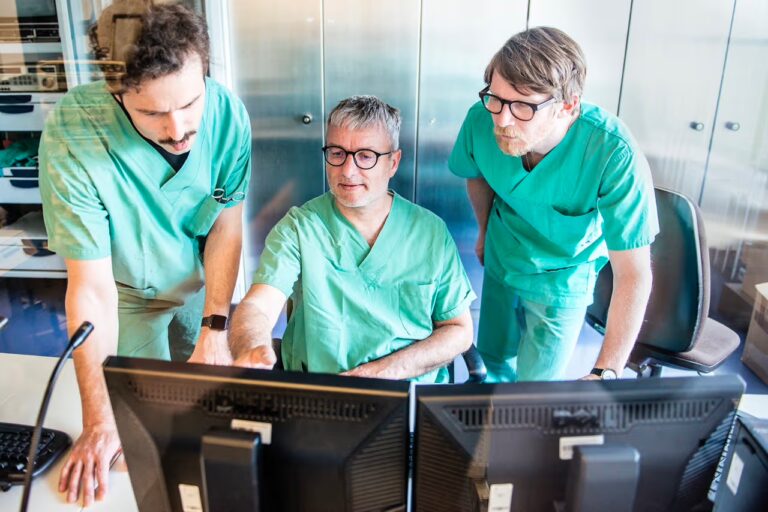 Drei medizinische Fachkräfte in grüner Arbeitskleidung arbeiten gemeinsam an einem Computer und besprechen Befunde in einem klinischen Arbeitsraum.