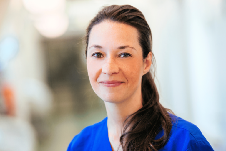 Porträt einer Frau mit zurückgebundenen Haaren und freundlichem Ausdruck vor einem unscharfen Hintergrund.