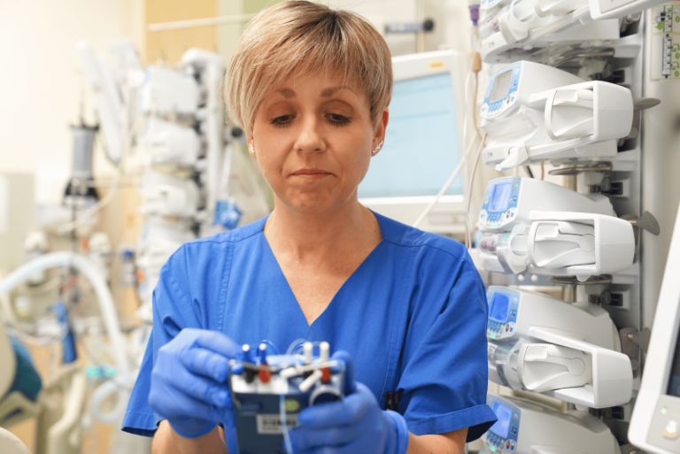 Medizinische Fachkraft in blauer Arbeitskleidung arbeitet in einer Klinik an einem technischen Gerät; im Hintergrund sind medizinische Geräte und Monitore zu sehen.