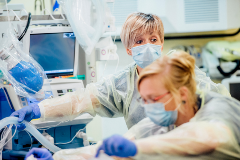 Zwei medizinische Fachkräfte mit Schutzkleidung und Masken arbeiten gemeinsam an einem Patientenarbeitsplatz mit medizinischen Geräten in einer Klinik.