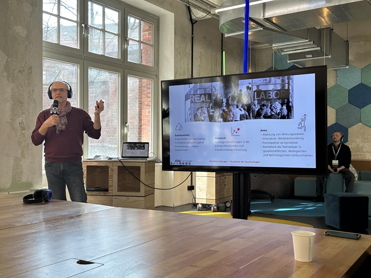 Ein Referent steht in einem modernen Veranstaltungsraum vor einem Bildschirm und präsentiert Inhalte zum Thema urbane Innovation und Reallabore; im Hintergrund sind Teilnehmende und eine Projektion mit Präsentationsfolien zu sehen.