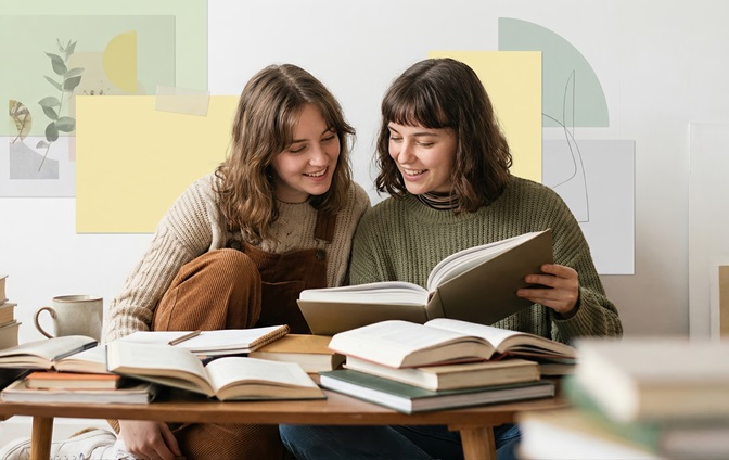 Zwei junge Frauen sitzen gemeinsam am Tisch, lernen und schauen in ein aufgeschlagenes Buch, umgeben von weiteren Lernmaterialien.