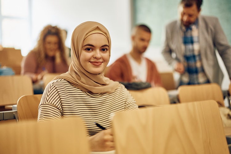 Junge Frau mit Kopftuch sitzt in einem Seminarraum und lächelt in die Kamera, während im Hintergrund weitere Studierende an Tischen sitzen.