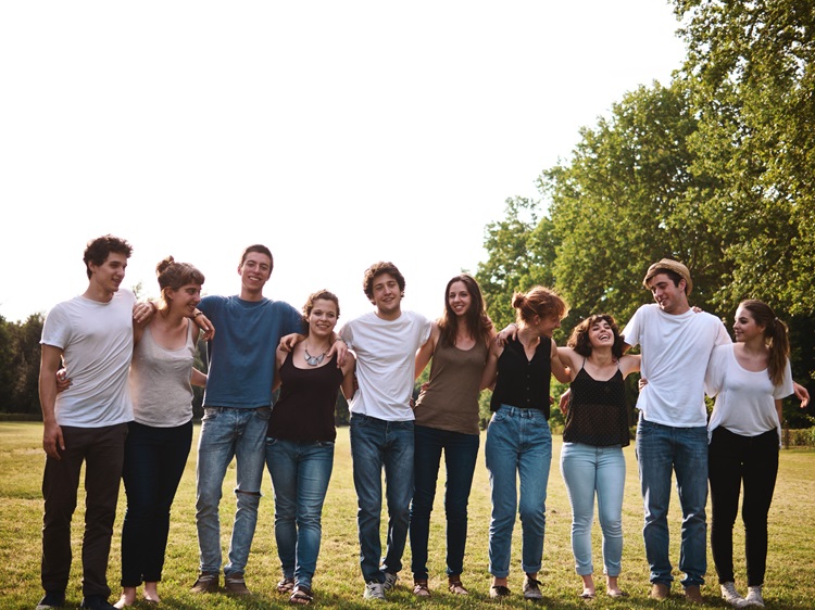 Mehrere junge Menschen stehen im Freien in einer Reihe, lächeln und wirken vertraut miteinander, im Hintergrund sind Bäume und eine natürliche Umgebung zu sehen.