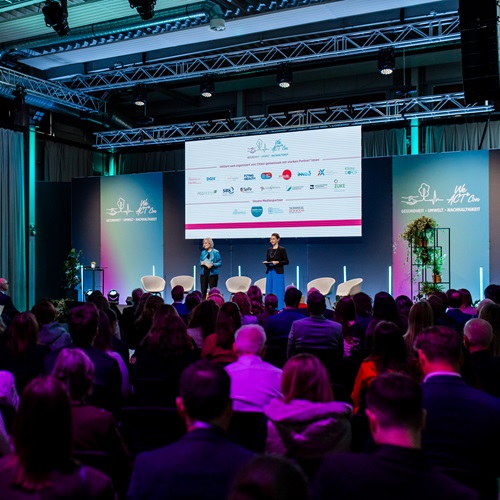 Publikum bei einer Konferenzveranstaltung in einem Saal mit Bühne und großer Präsentationsleinwand; Vortragssituation mit mehreren Referierenden vor beleuchtetem Hintergrund.