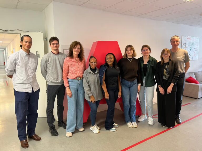 Mehrere internationale Teilnehmende und Mitarbeitende des IRIA stehen gemeinsam in den Räumlichkeiten der Akkon Hochschule und posieren für ein Gruppenfoto vor einer roten Wand.