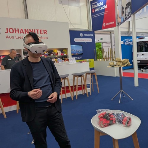 Ein Mann mit VR-Brille steht an einem Messestand der Johanniter und testet eine virtuelle Anwendung. Im Vordergrund befindet sich ein Stehtisch mit mehreren VR-Controllern. Im Hintergrund sind Informationsstände und Sitzmöglichkeiten der Messe zu sehen.