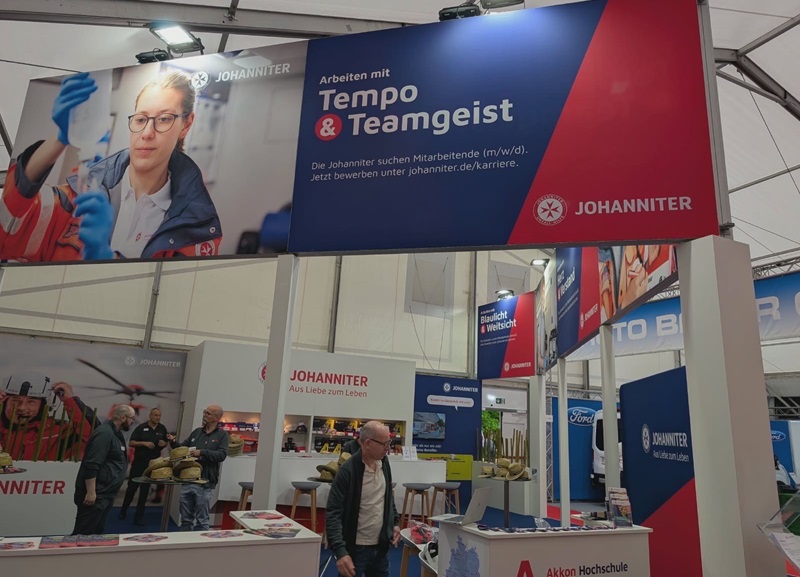 Messestand der Johanniter auf einer Fachmesse mit großem Banner „Tempo Teamgeist“. Im Hintergrund befinden sich Besucherinnen und Besucher sowie verschiedene Ausstellungsstände der Messehalle.