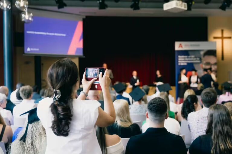 Absolvent*innen der Akkon Hochschule werfen während der Abschlussfeier ihre Hüte in die Luft und feiern gemeinsam ihren erfolgreichen Studienabschluss. Im Vordergrund verfolgen Gäste die Zeremonie, während auf der Bühne die Urkundenverleihung stattfindet.