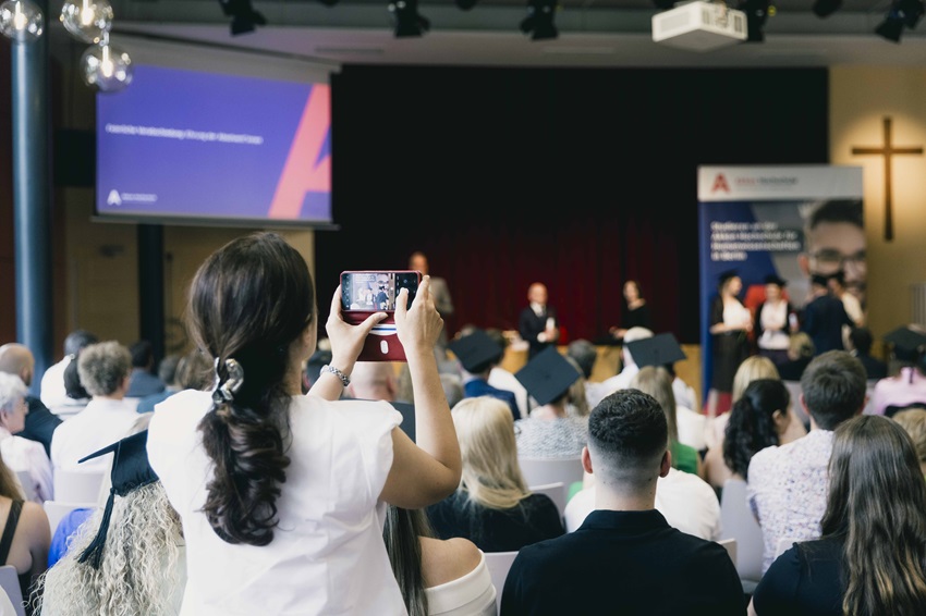 Absolvent*innen der Akkon Hochschule werfen während der Abschlussfeier ihre Hüte in die Luft und feiern gemeinsam ihren erfolgreichen Studienabschluss. Im Vordergrund verfolgen Gäste die Zeremonie, während auf der Bühne die Urkundenverleihung stattfindet.