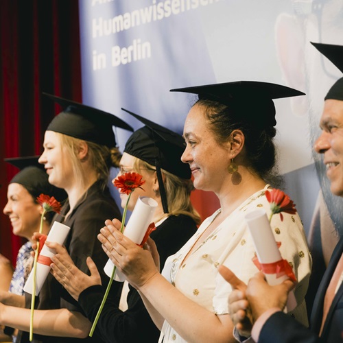 Studierende der Akkon Hochschule tragen bei einer Abschlussfeier traditionelle Absolvent*innenhüte und freuen sich über ihren erfolgreichen Studienabschluss. Im Hintergrund sind weitere Teilnehmende der Feier zu sehen.