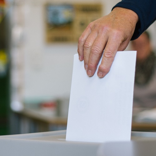 Eine Person wirft einen weißen Stimmzettel in eine Wahlurne. Der Fokus liegt auf der Hand und dem Stimmzettel, der Hintergrund ist unscharf.