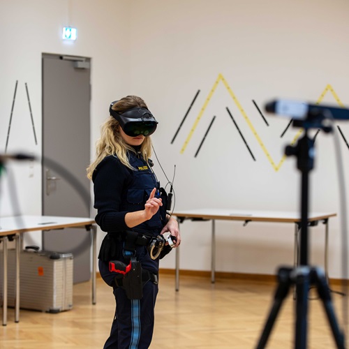 Eine Person trägt eine Virtual-Reality-Brille und Sensoren an Händen und Körper, während sie in einer Forschungsumgebung eine VR-Anwendung testet. Die Aufnahme veranschaulicht den Einsatz von Virtual-Reality-Technologien zur Erforschung und zum Training kommunikativer Deeskalation im Forschungsprojekt K3VR.
