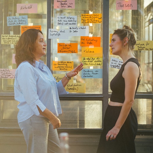 Zwei Frauen stehen in einem modernen Hochschulgebäude vor einer Glaswand mit zahlreichen farbigen Haftnotizen und tauschen sich über Projektergebnisse oder Ideen aus. Die Szene vermittelt einen praxisnahen und kollaborativen Arbeitsprozess im Hochschul- oder Forschungskontext.