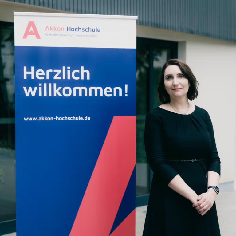 Andrea Ferber, Geschäftsführerin der Akkon Hochschule, steht neben einem Willkommensbanner im Eingangsbereich der Hochschule und blickt in die Kamera.