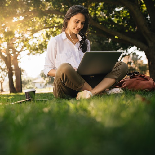 Studentin sitzt mit einem Laptop auf einer Wiese im Park und lernt im Freien. Die sonnige Atmosphäre steht für flexibles, ortsunabhängiges Studium und modernes Lernen.