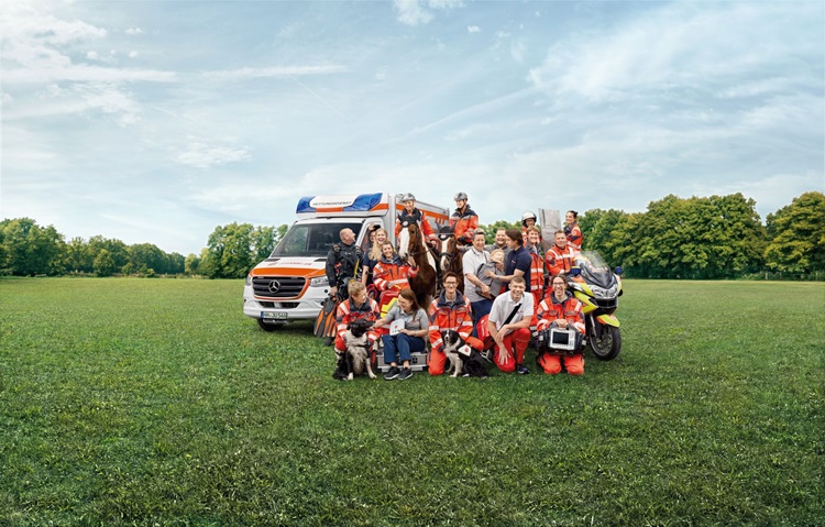Gruppenfoto eines Teams auf einer Wiese. Die Teilnehmenden sitzen und stehen gemeinsam vor einem bewölkten Himmel und blicken in die Kamera.